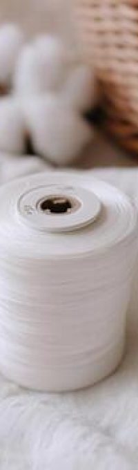 a-white-spool-of-thread-sits-on-a-textured-white-fabric-with-cotton-bolls-and-a-wicker-basket-in-the-soft-focus-background-photo